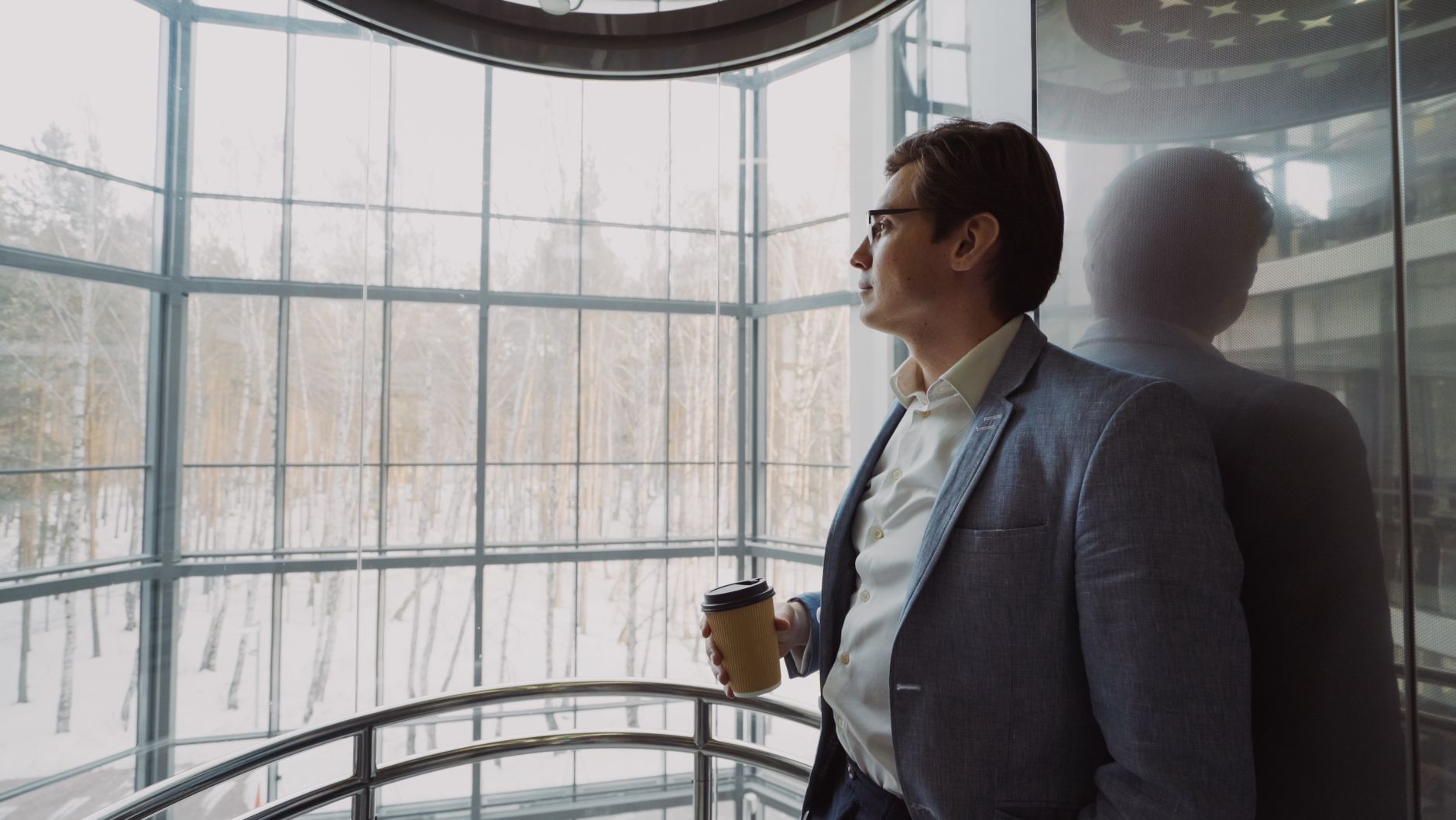 Businessman in blue suit with coffee cup looking out a large window, reflecting on a winter landscape. — Optimize Connectivit